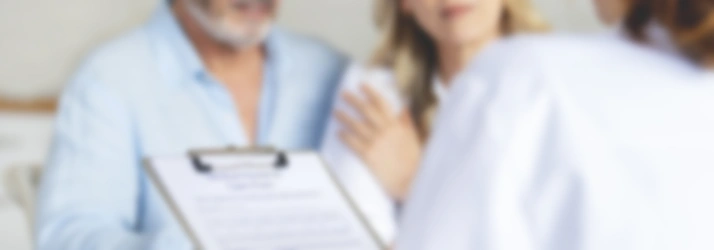 A healthcare professional holds a clipboard with a form while speaking with two individuals seated nearby